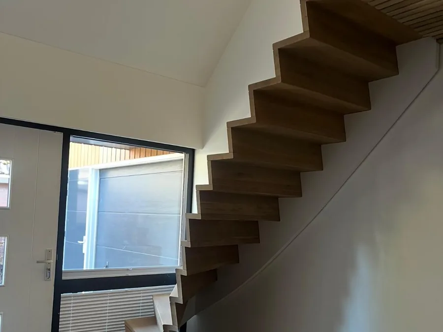 Hal van een nieuwbouwwoning met een houten zwevende trap langs een witte wand